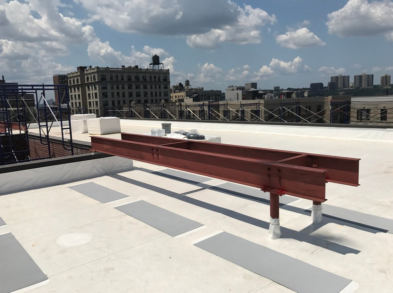 Red steel beams on a flat rooftop in an urban setting, with buildings and a water tower visible under a blue sky.