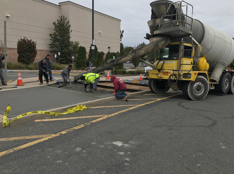 Construction crew pouring concrete in a parking lot; yellow cement truck, caution tape, workers.