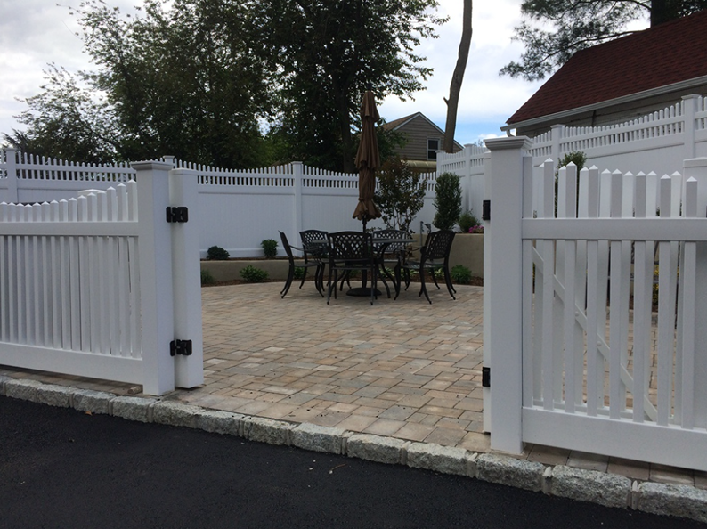 White picket fence with open gate leading to brick patio with table and chairs.