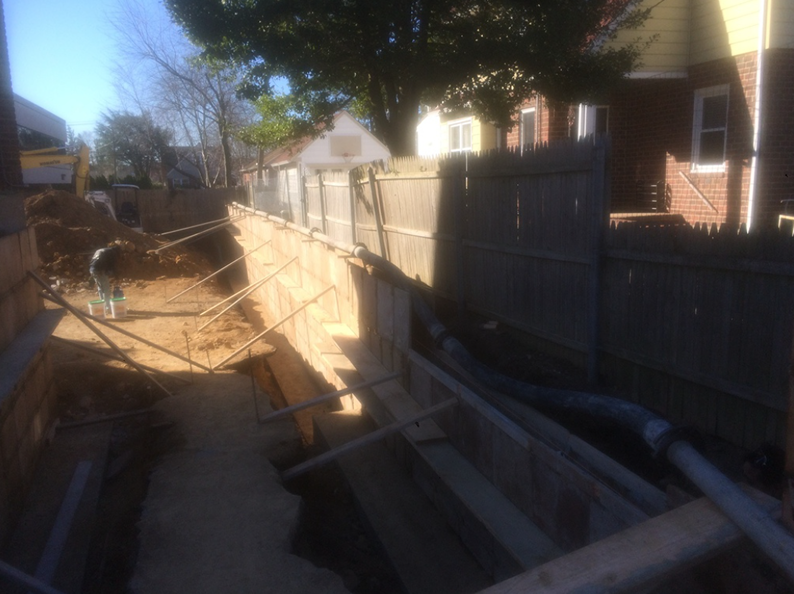 Construction site, trench dug along wooden fence, with support beams and pipes.