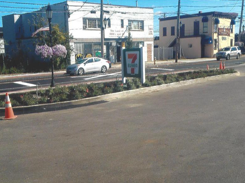 Street view: A 7-Eleven sign, road with a median, car, buildings, and a traffic cone.