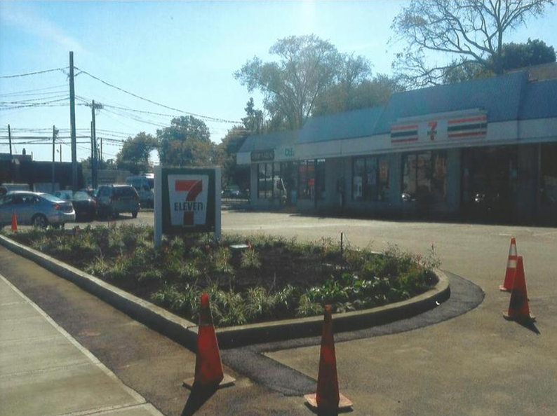 A 7-Eleven store with a sign and surrounding landscaped area. Traffic cones are on the road in front.