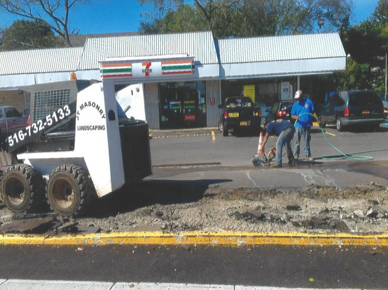Construction in front of 7-Eleven: skid steer, workers cutting asphalt, parked vehicles, blue sky.