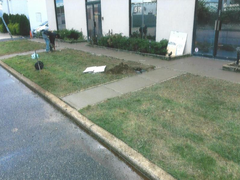 Sidewalk construction: Person pouring concrete near a building entrance, surrounded by grass and curb.