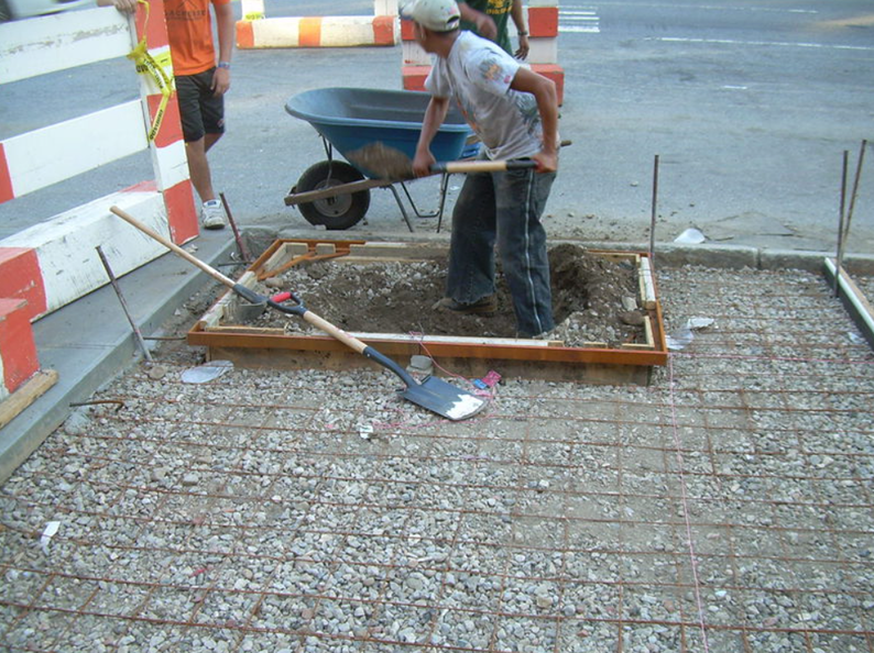 Construction workers digging in a framed area on a concrete surface with a wheelbarrow, tools, and barriers.