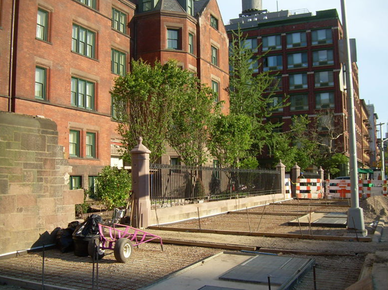 Construction site with gravel and metal, next to a brick building and a wrought iron fence.