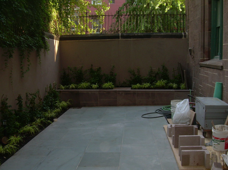 A small, stone patio with a built-in planter. Plants line the walls of the space. Construction materials present.