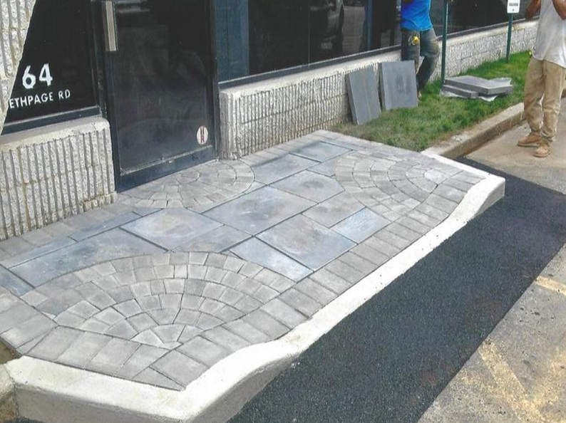 Entryway with patterned pavers, asphalt border, and a concrete curb. A person stands nearby.