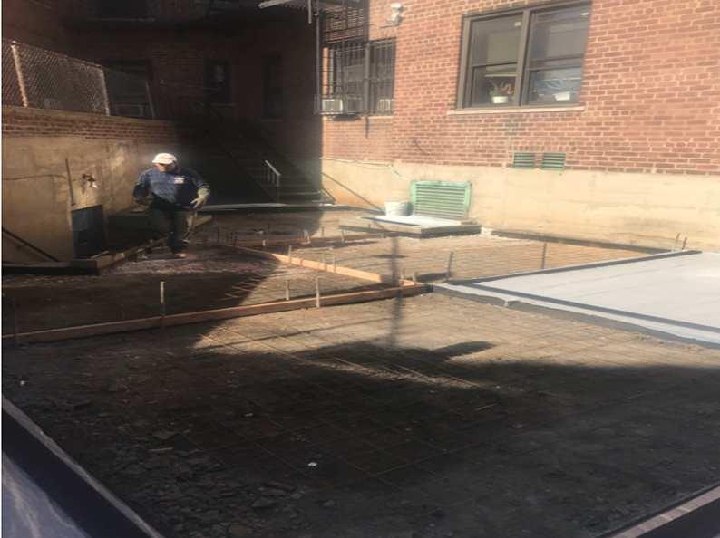 Worker smoothing wet concrete in a construction area, with wooden forms and brick building.