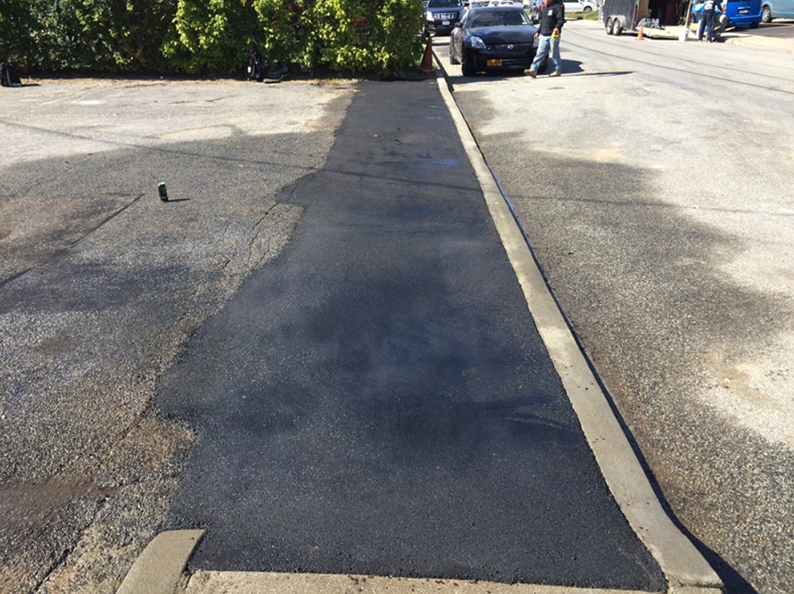 Freshly paved asphalt pathway next to a curb and bushes, with a car and people in the background.
