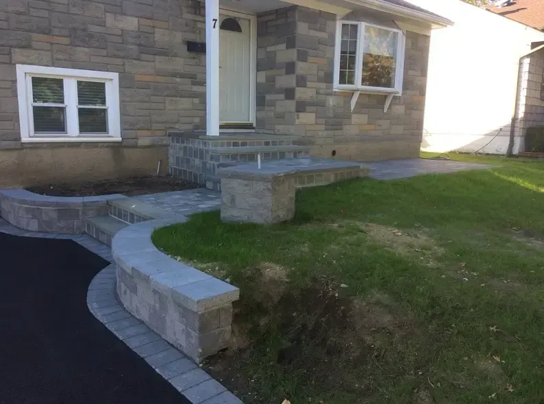 Stone house with steps, walkway, and retaining walls; green lawn on right, asphalt on left.