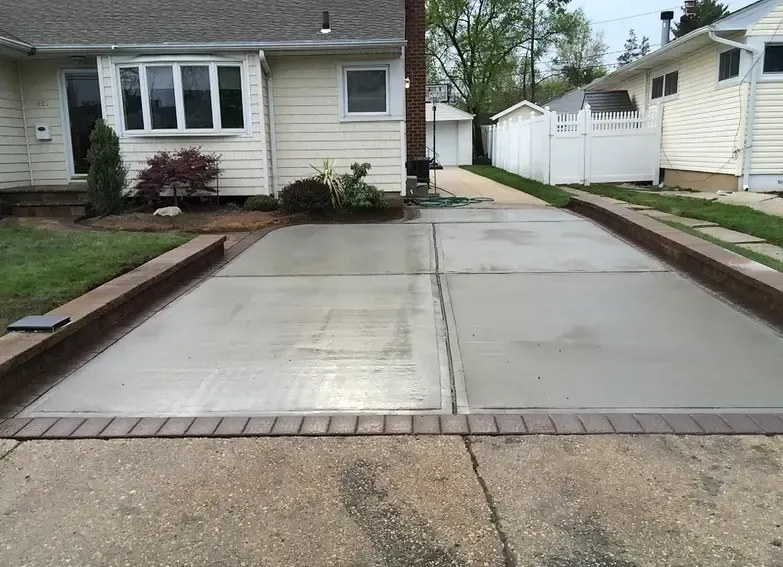 Newly poured concrete driveway bordered by brick, in front of a light-colored house.