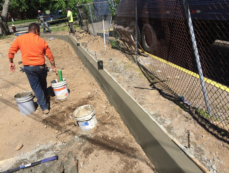 Construction worker in orange shirt carrying buckets; a concrete curb is being built on a sidewalk.