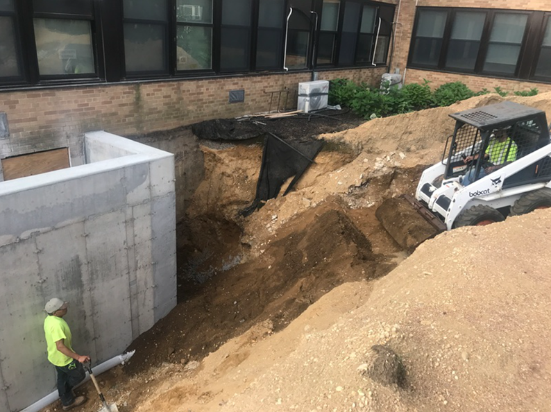 Construction site with excavator digging next to a building with a concrete foundation wall. A worker shovels dirt.