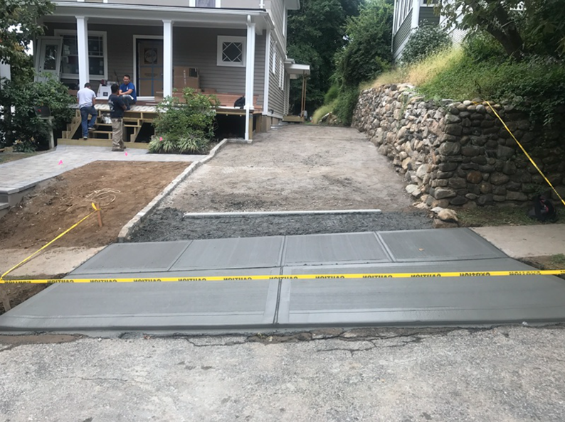 Concrete driveway construction in progress; workers, house, retaining wall, caution tape, gray and brown.