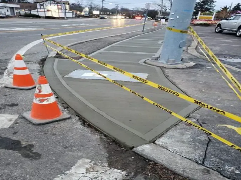 Freshly poured concrete curb with caution tape and cones near a road.