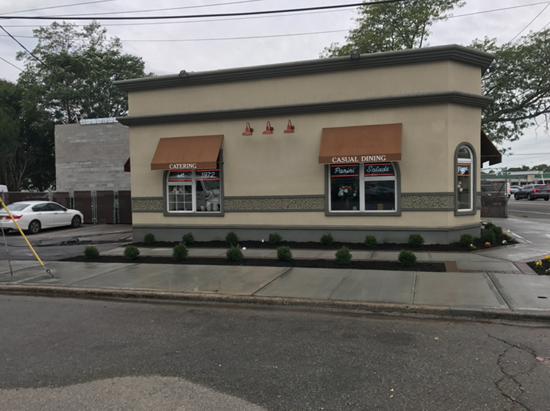 Tan building with brown awnings, storefronts, and small shrubs lining the sidewalk in front.