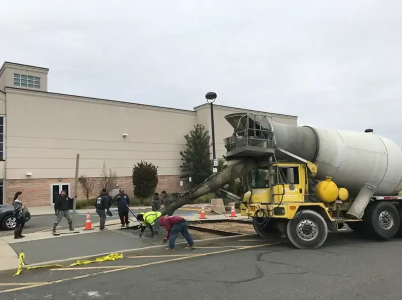 Yellow cement truck pouring concrete into a sidewalk repair area, workers assisting, near a building.