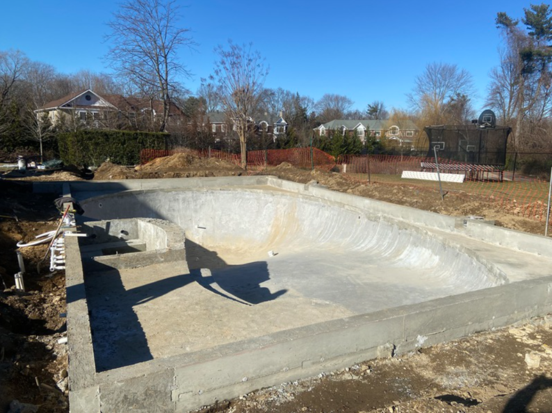 Concrete pool under construction in a yard on a sunny day. Houses and trees in the background.