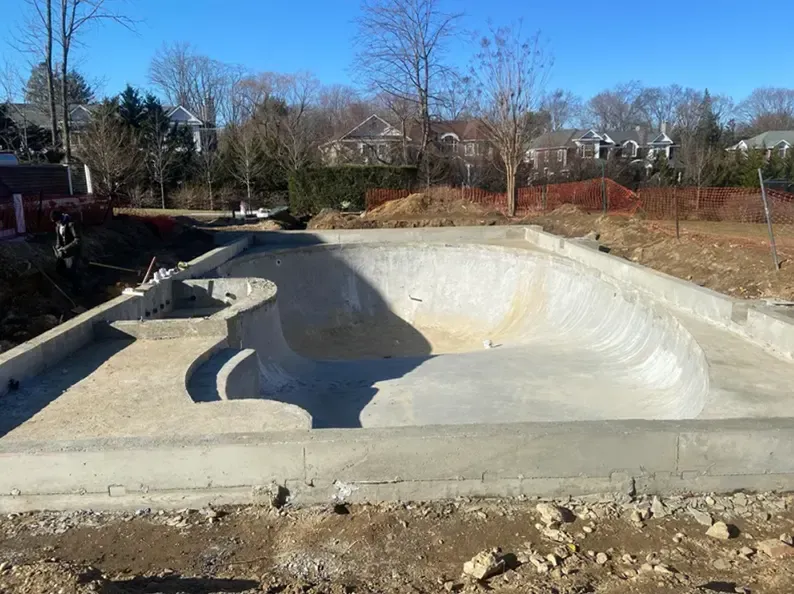 Construction of a concrete skate bowl; exterior shot with dirt and houses in the background.