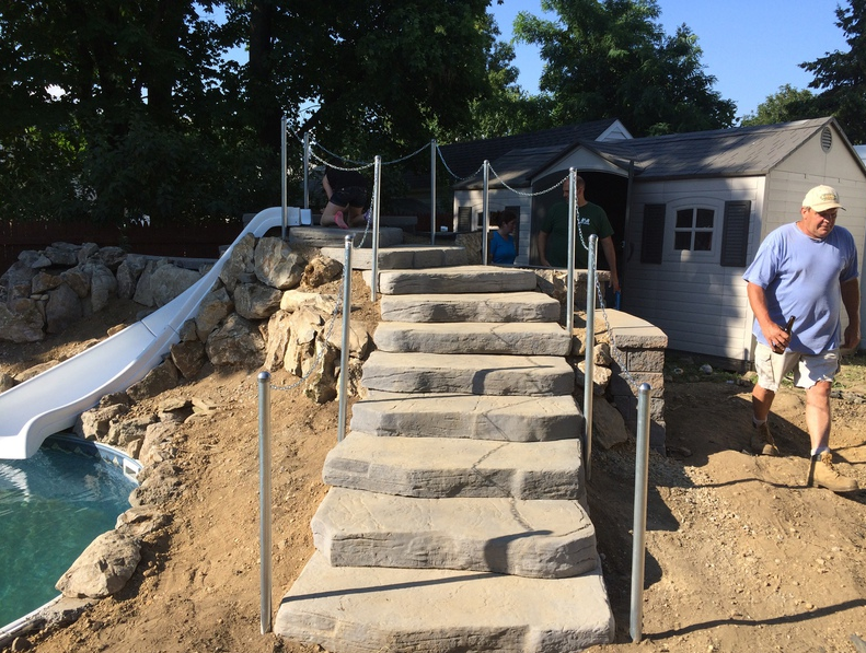 Stone stairs lead to a pool slide and playhouse. Man walks towards stairs, holding something.