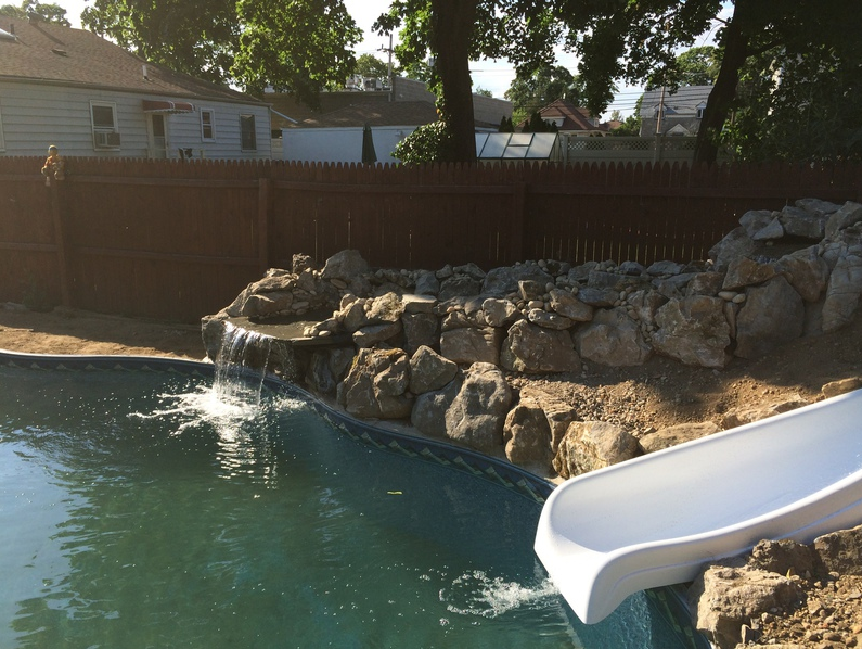 Pool with rock waterfall and white slide; brown fence and houses in background.