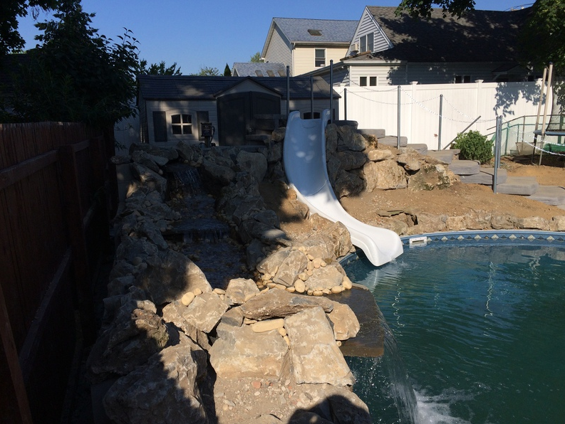 Backyard pool with rock waterfall, white slide, and wooden fence.