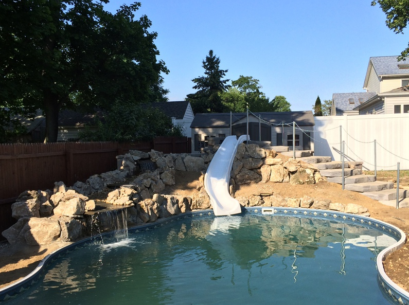 Swimming pool with a white slide, waterfall, and stone features in a backyard.