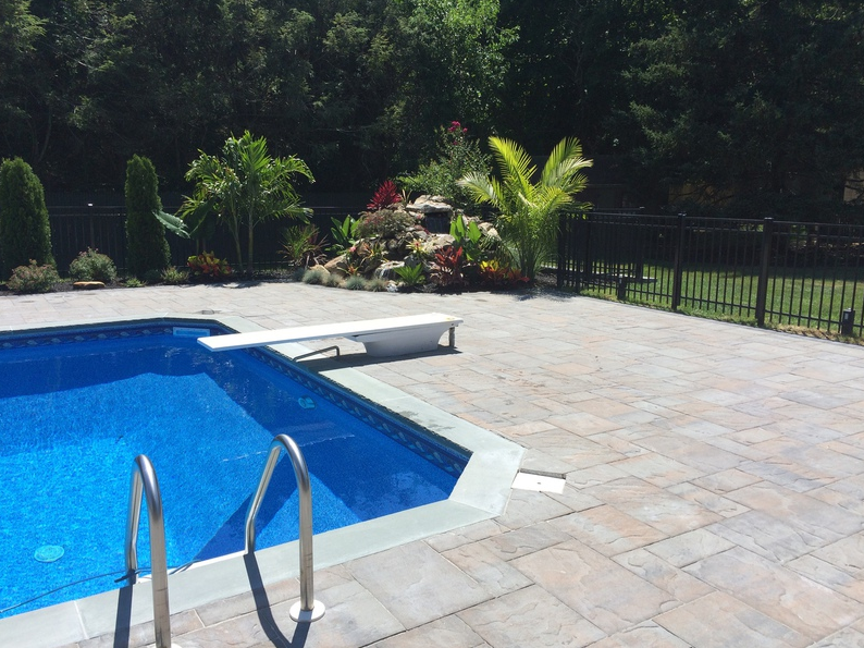 Swimming pool with diving board, brick patio, and landscaped area against a black fence.