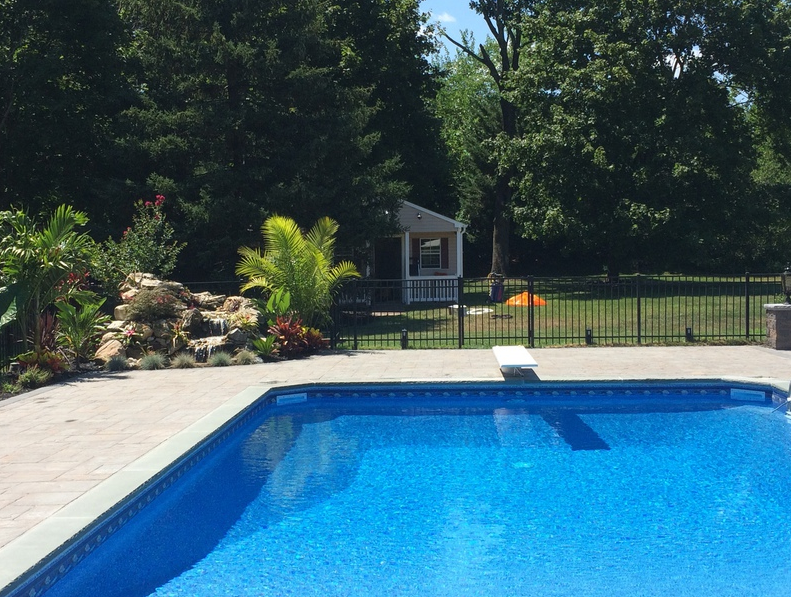 Backyard pool with blue water, diving board, landscaping, and fence, under a blue sky.