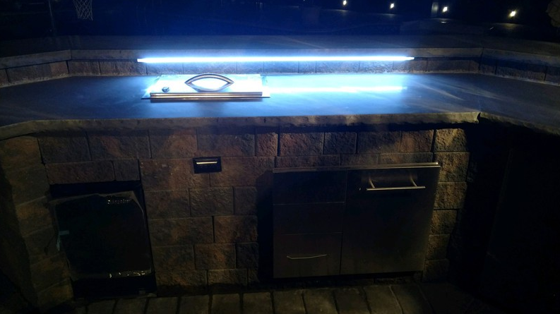 Outdoor kitchen with stone facade, lit countertop, grill, and storage cabinets.