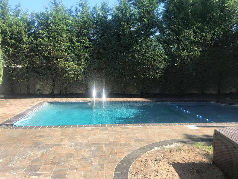 A swimming pool with fountain jets, surrounded by pavers and evergreen trees.