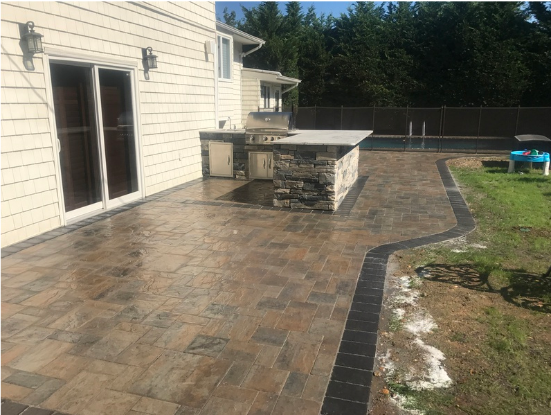 Patio with a built-in grill, leading to a pool. Paved with brown and black bricks next to a house.