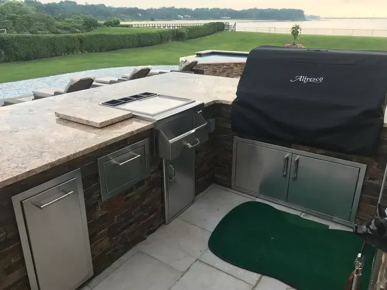 Outdoor kitchen with grill, storage, and putting green on a patio, overlooking water.