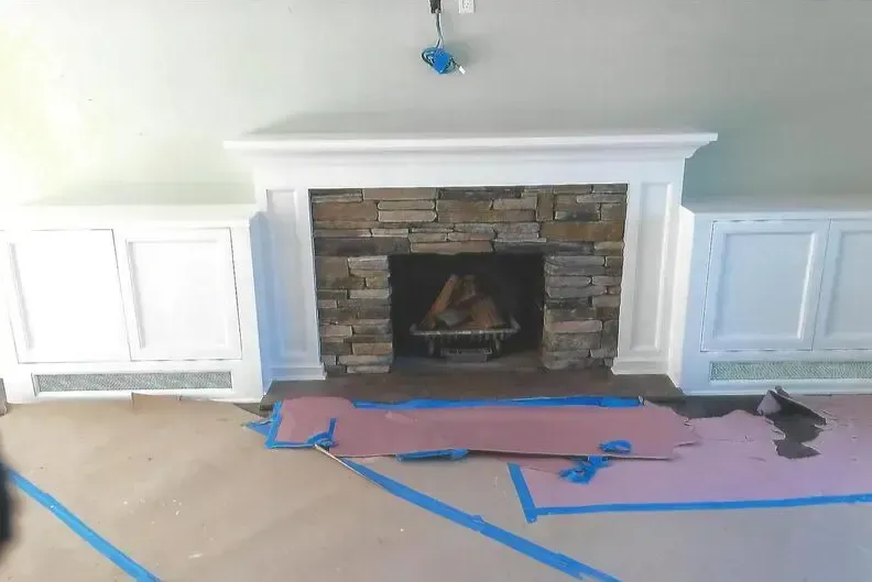 Fireplace with stone surround, white mantel and flanking cabinets. Room under construction, covered floor.