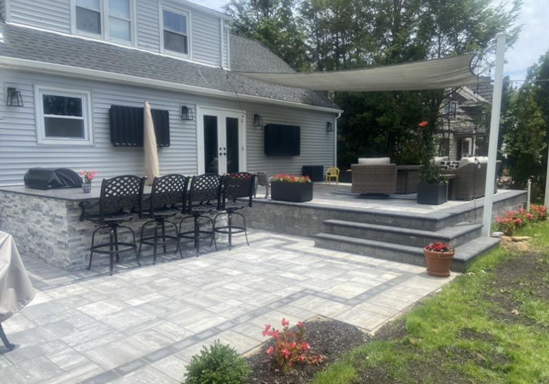 Backyard patio with bar seating, grill, shade sail, and steps leading to a raised outdoor living area.