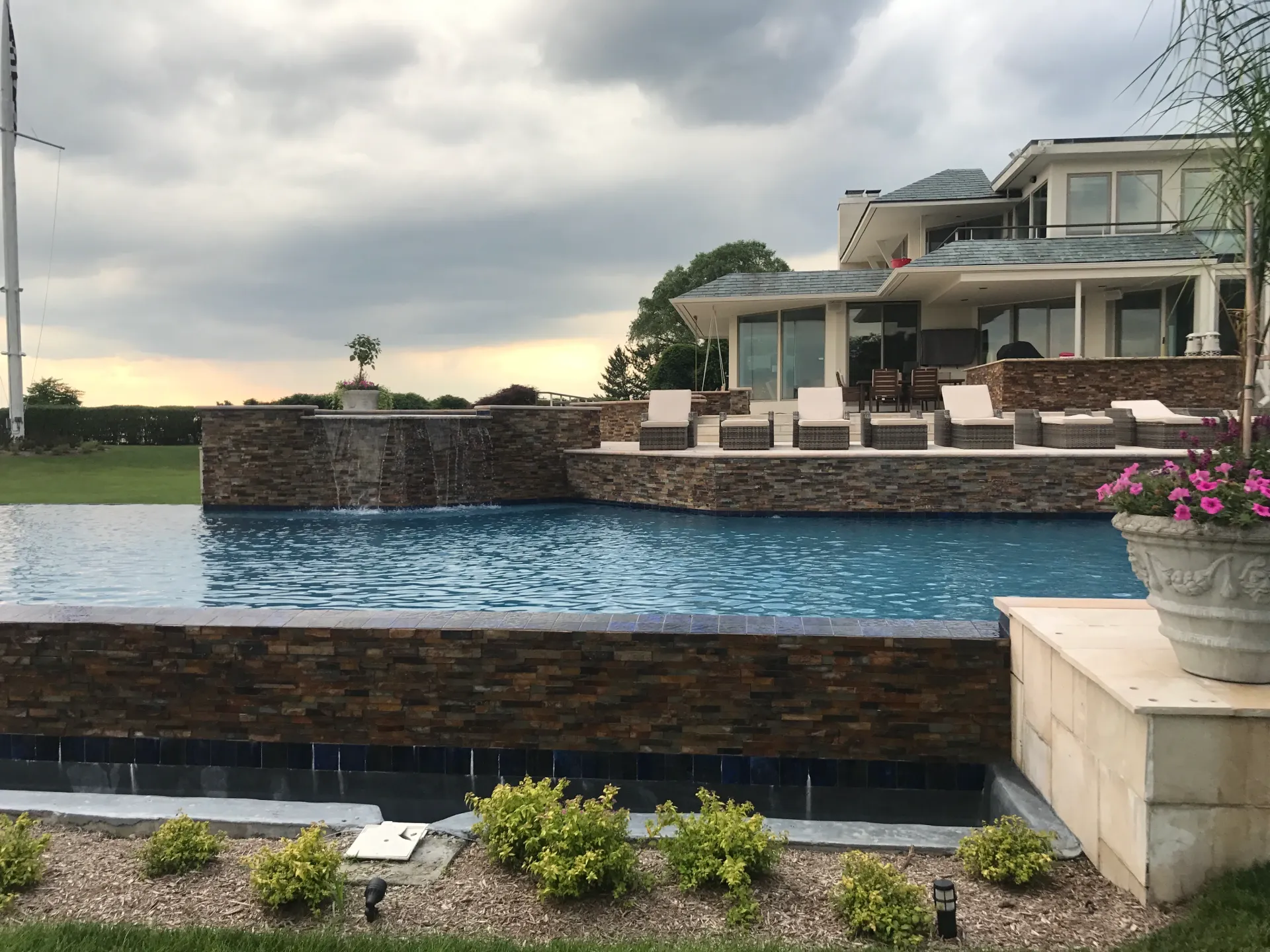 Luxurious backyard with infinity pool, lounge chairs, and modern house with stone accents under a cloudy sky.