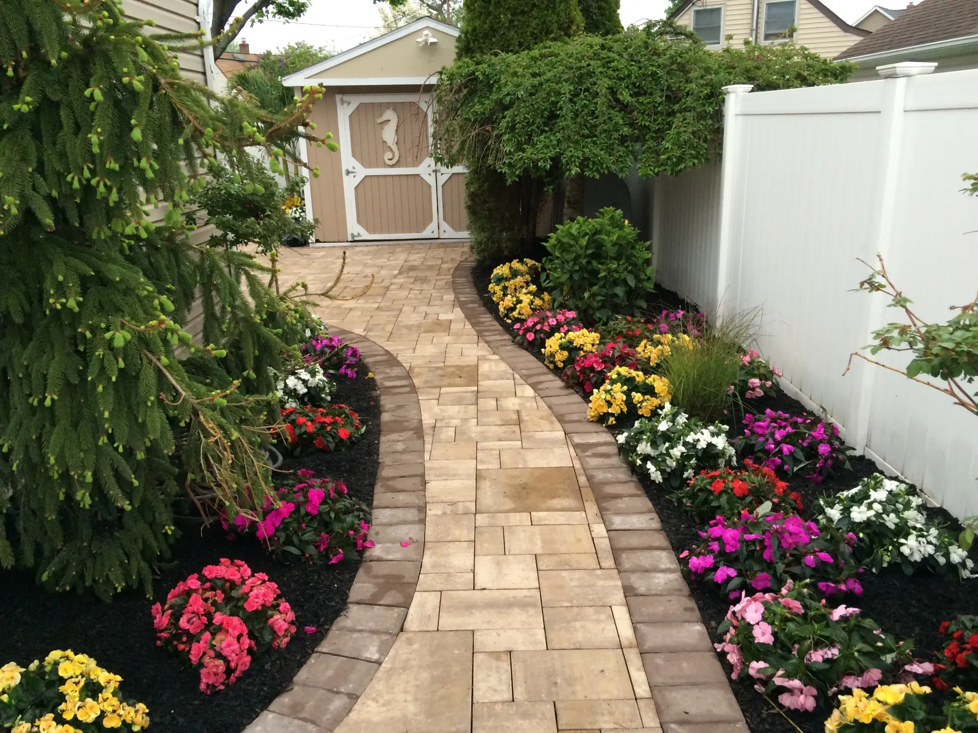 Stone pathway lined with colorful flowers, leading to a small shed with a seahorse design.