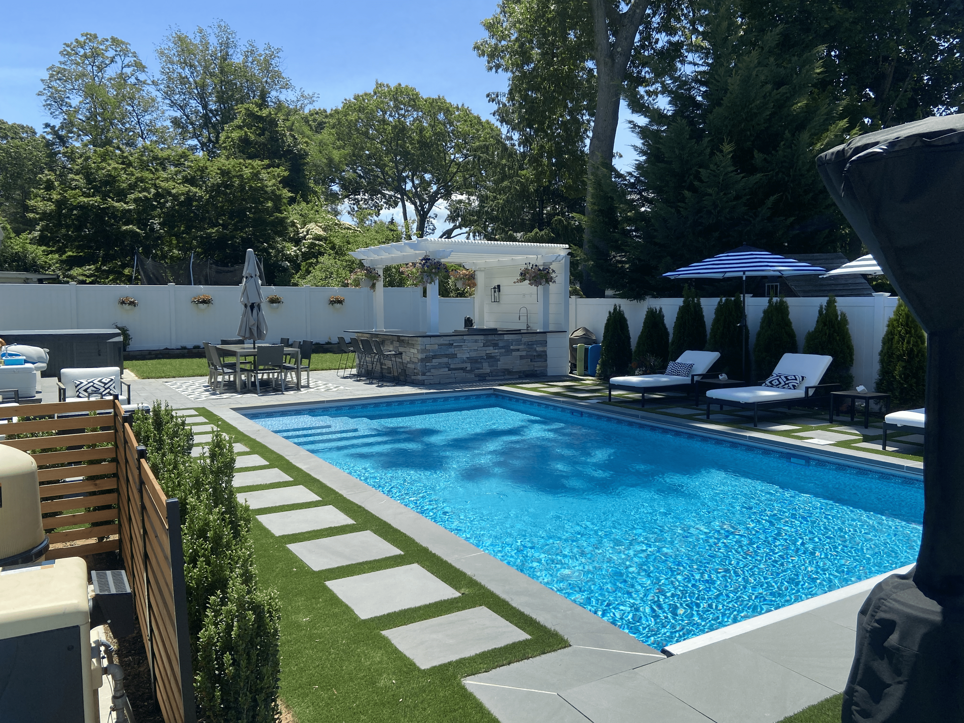 A backyard pool with stone patio and lounge chairs, under a blue umbrella, on a sunny day.