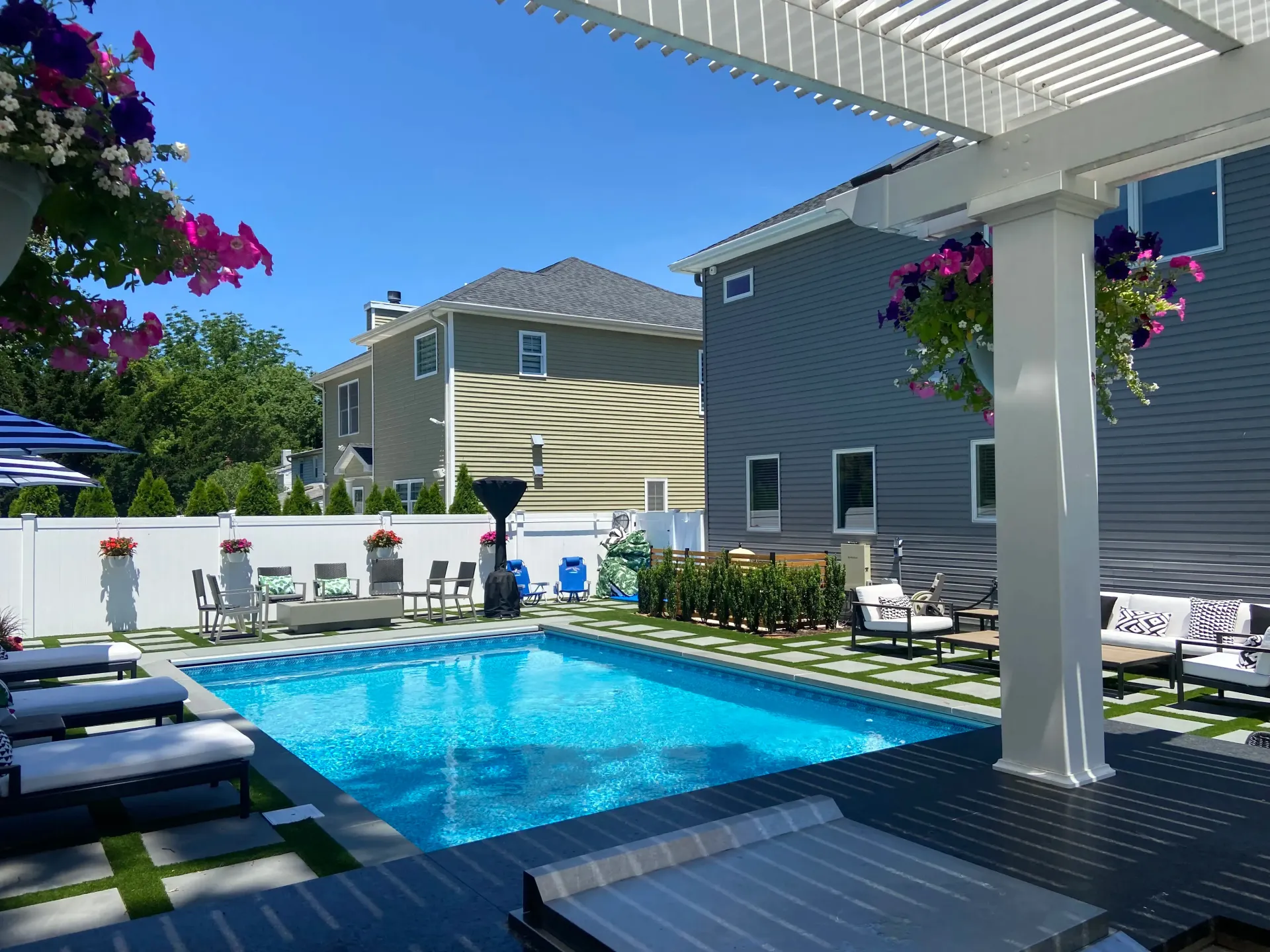 Pool area with blue water, flowers, patio furniture, and gray siding.