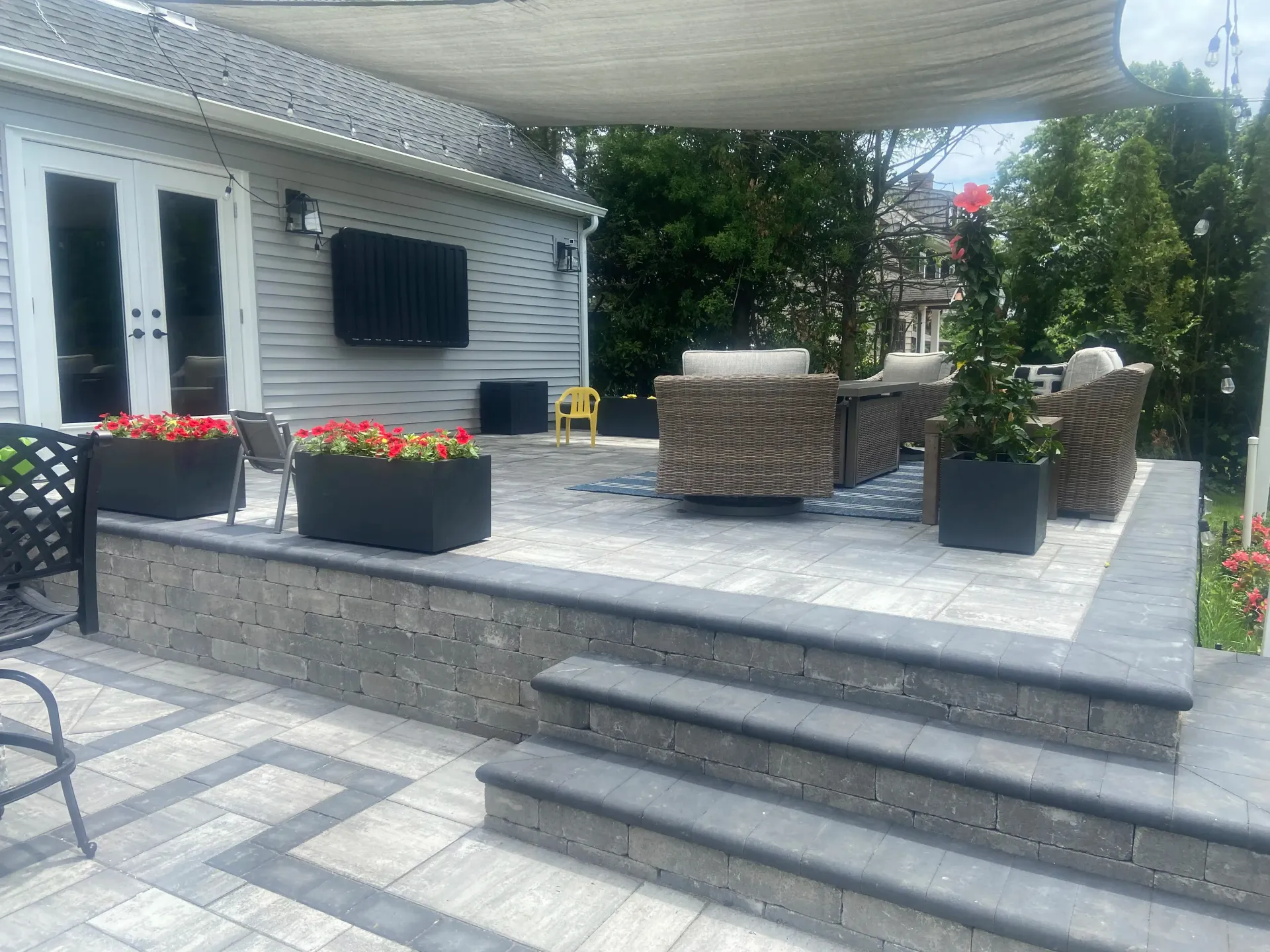 Backyard patio with seating, flower boxes, and steps. Gray pavers and stone wall. Shaded by a beige canopy.