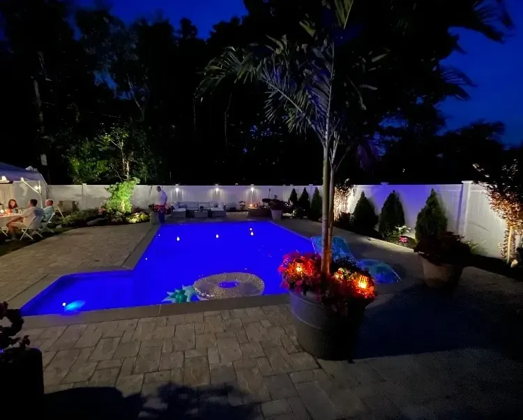 Pool at night with blue water, surrounded by a paved patio, illuminated by lights, with trees and plants.