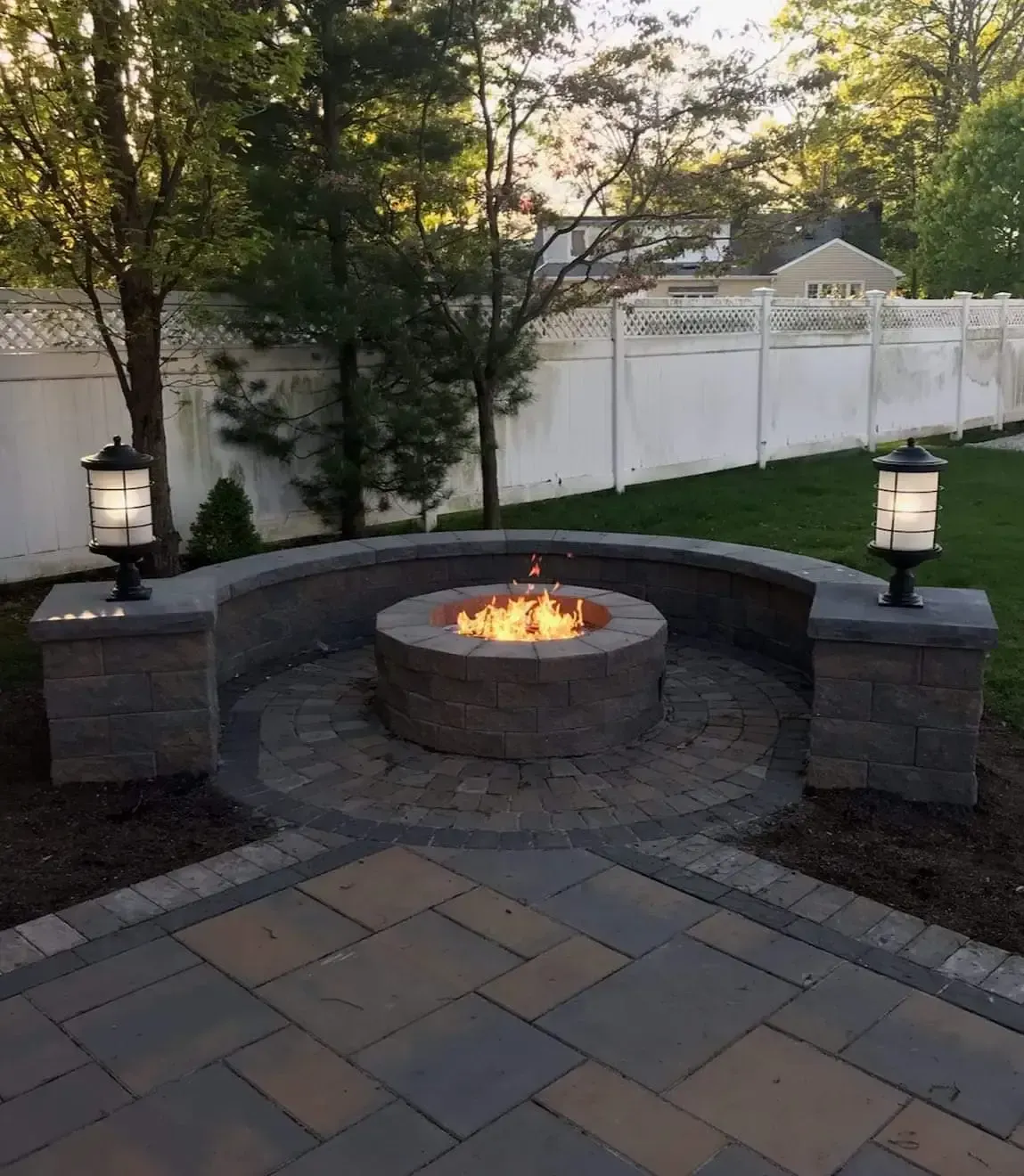 A backyard fire pit surrounded by a curved wall with built-in seating. Lighting and brick patio included.