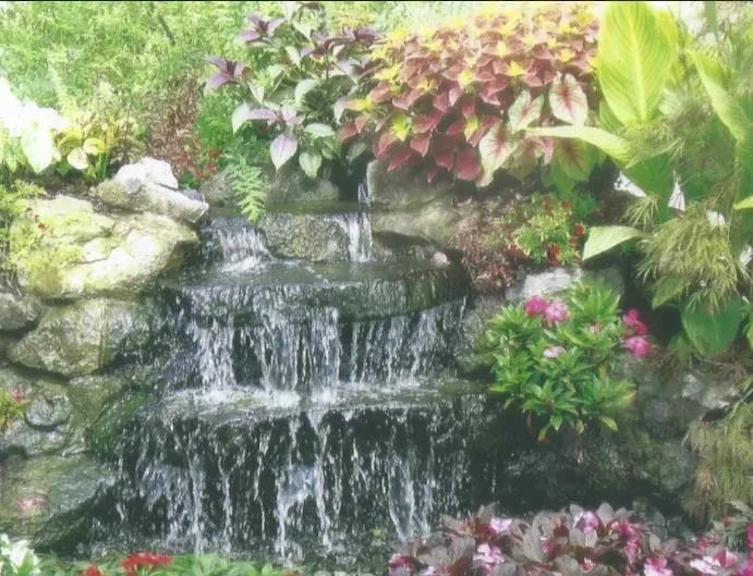 Small waterfall cascading down stone wall, surrounded by colorful flowers and foliage.