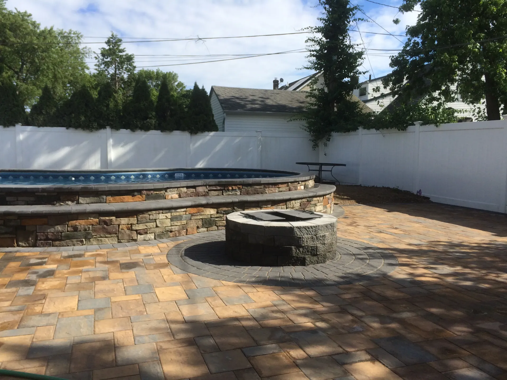 Backyard patio with a pool, fire pit, and paver bricks, surrounded by a white fence.
