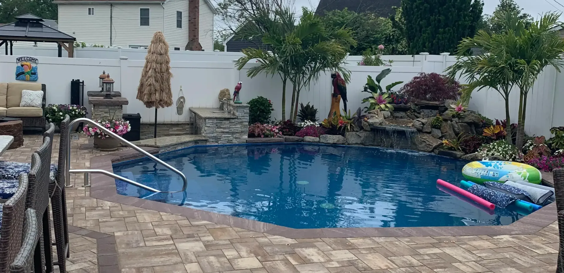 A backyard pool with a waterfall, patio furniture, and a white fence.