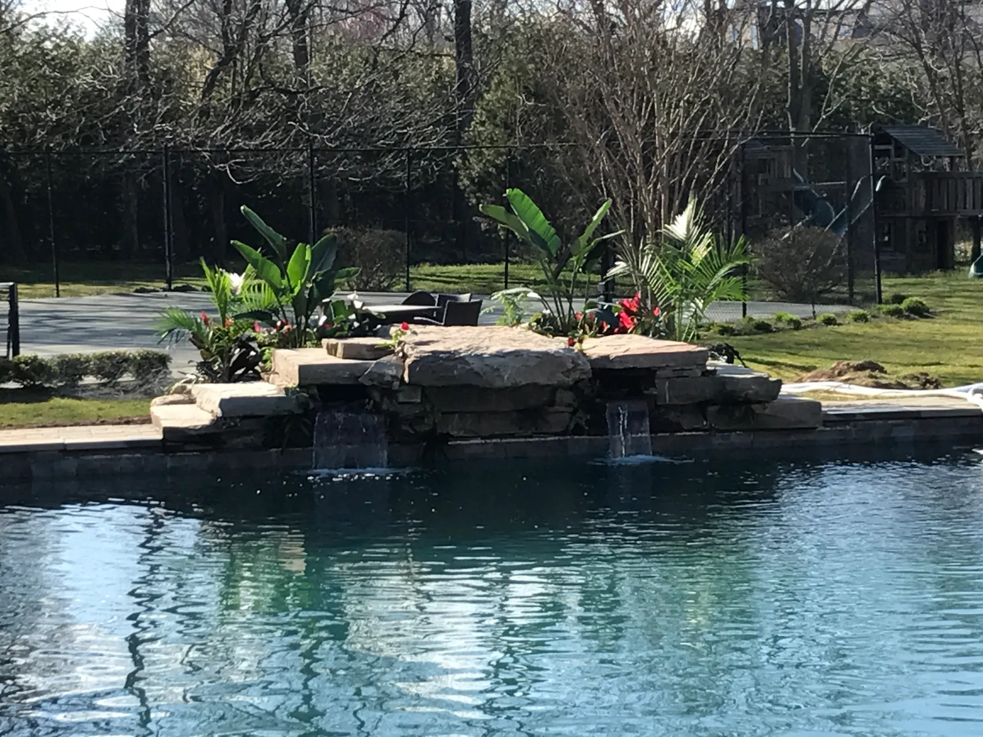 Pool with stone waterfall, plants, and a black fence in a backyard setting.