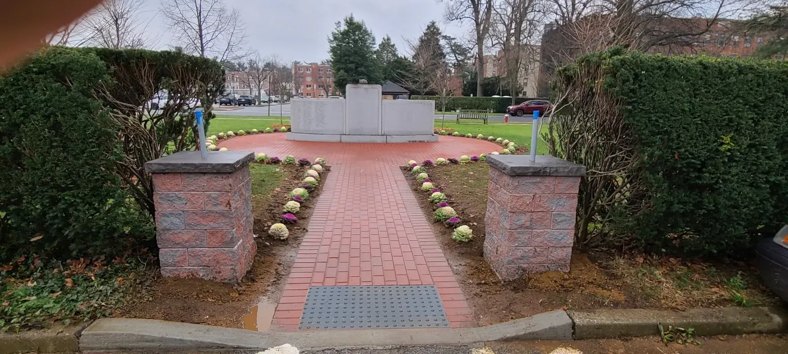 A brick pathway leads to a monument flanked by green hedges and flowerbeds on a cloudy day.