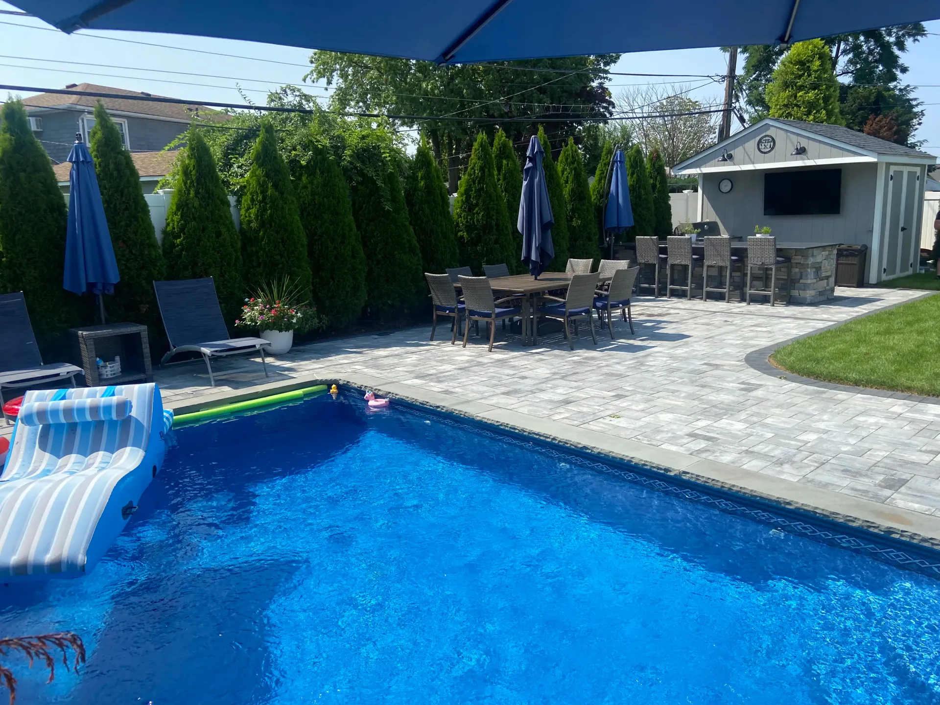 Pool area with blue water, lounge chairs, patio table, and outdoor kitchen under a blue umbrella.