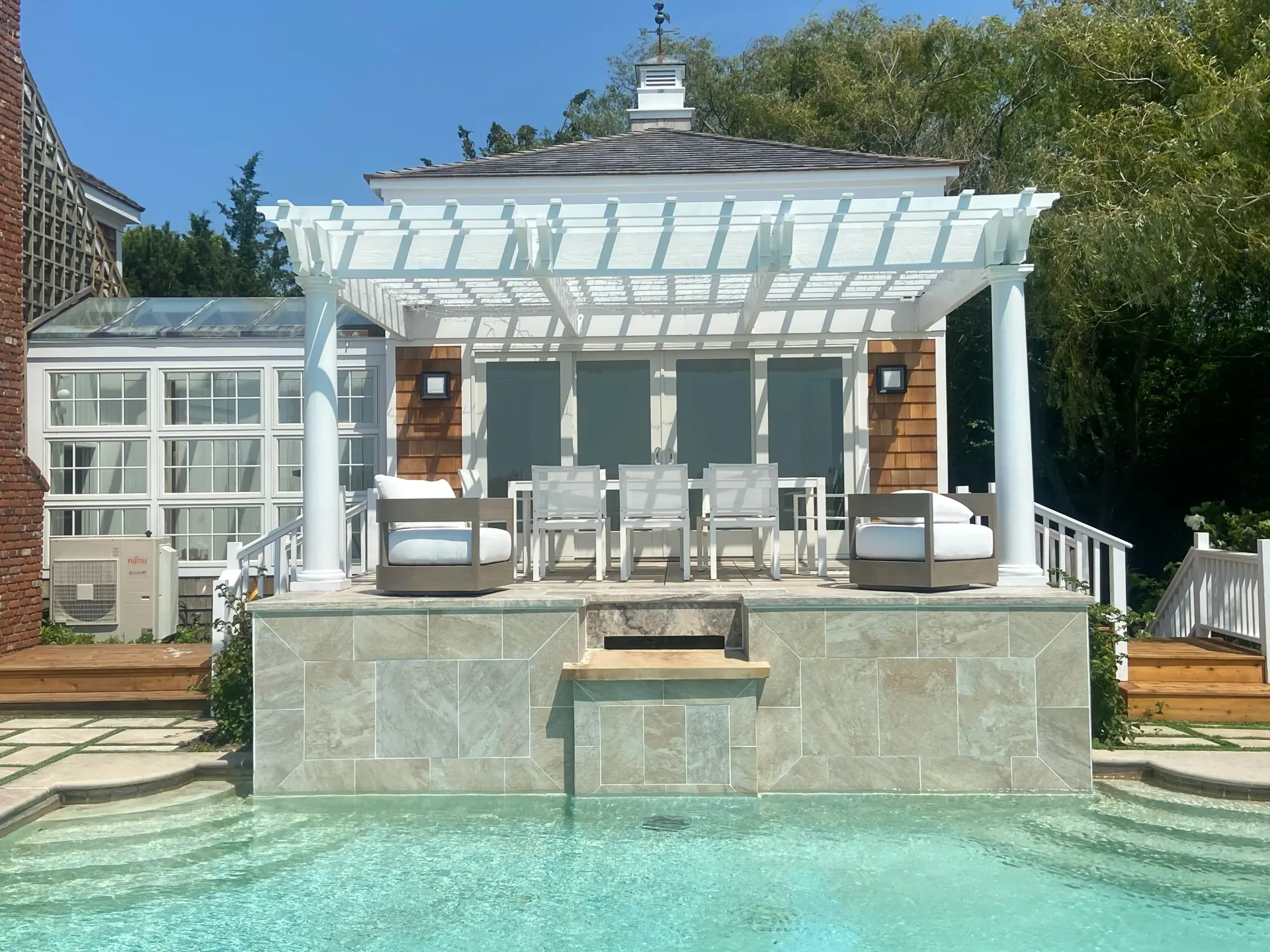 Poolside dining area with pergola and seating, overlooking the pool, with a blue sky.